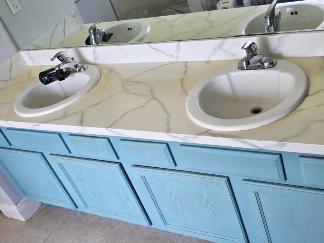 Painted faux-marble double vanity top, yellowed on the flat surface, with two drop-in sinks, blue cabinets