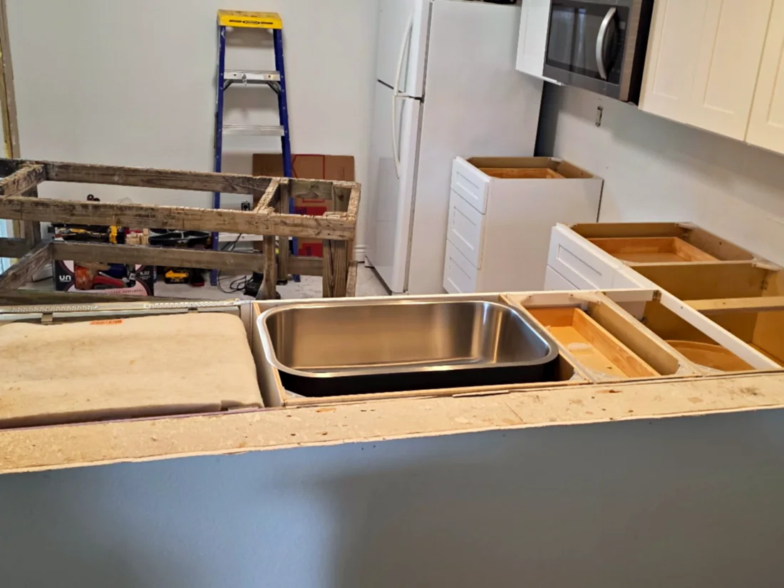 Unfinished kitchen pass-through with white shaker base cabinets, stainless undermount sink and raw particleboard edge